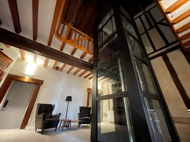 Salon de la maison avec la cage d’ascenseur vitrée et les poutres anciennes.