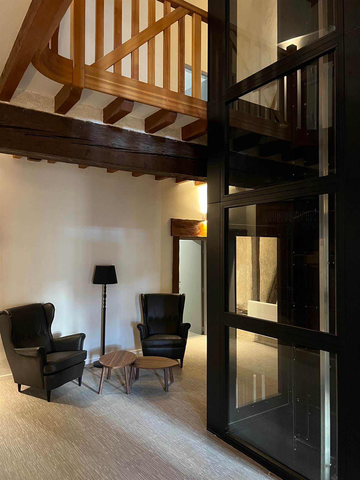 Palier avec deux fauteuils, une lampe, et la cage d’ascenseur vitrée noire traversant la charpente ancienne.
