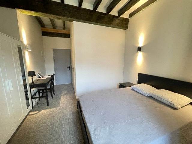 Chambre sous poutres avec un lit, un bureau devant la fenêtre et une armoire.