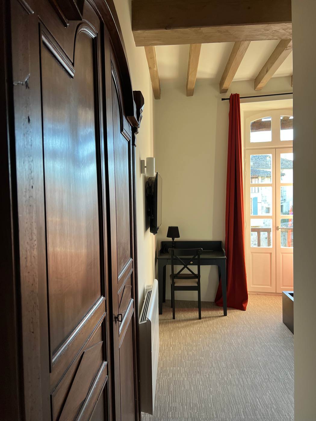 Chambre avec armoire ancienne, bureau devant la porte-fenêtre et rideau rouge.