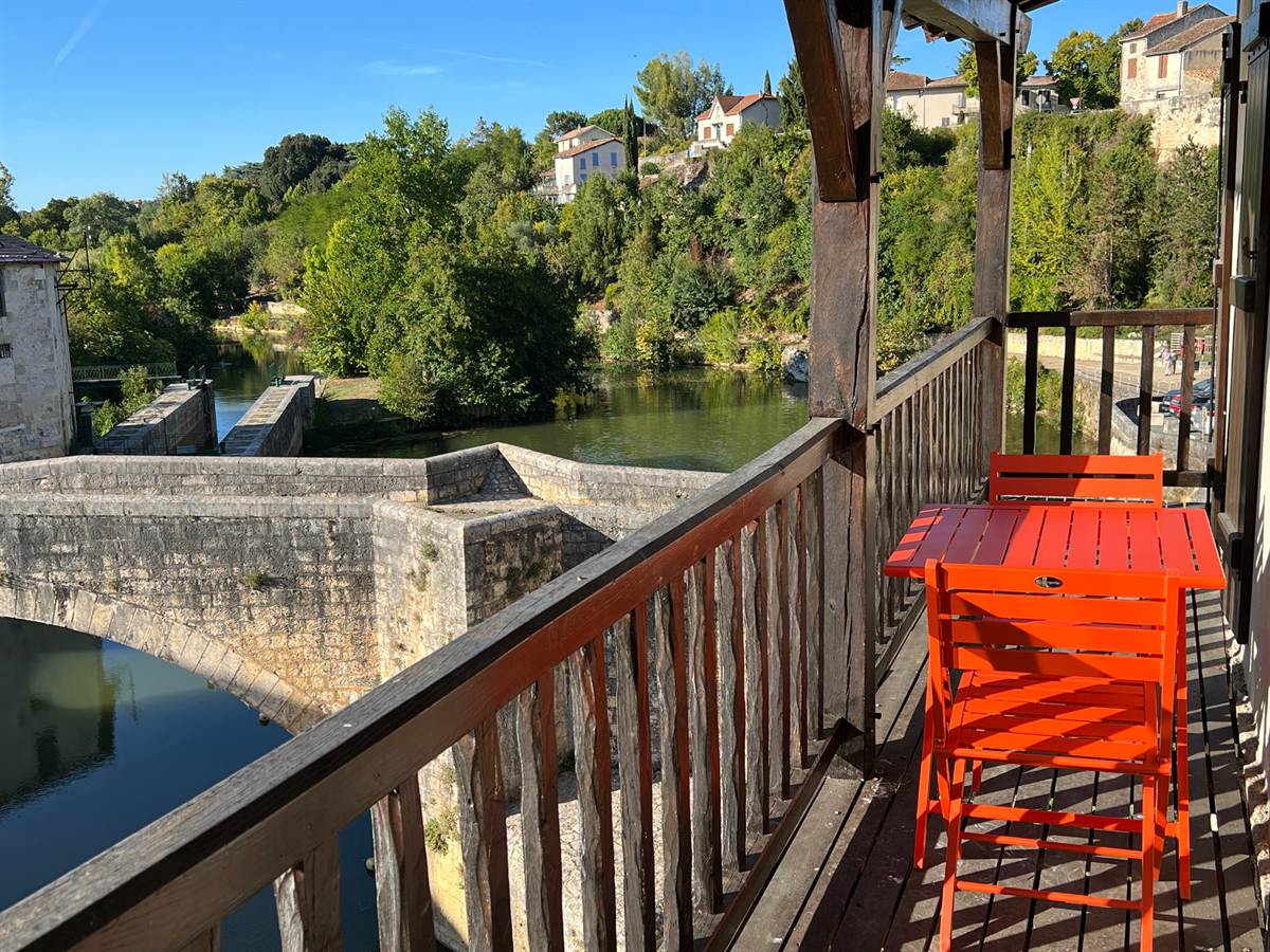 Balcon de bois avec une table et des chaises rouges, surplombant la Baïse et le pont de pierre.