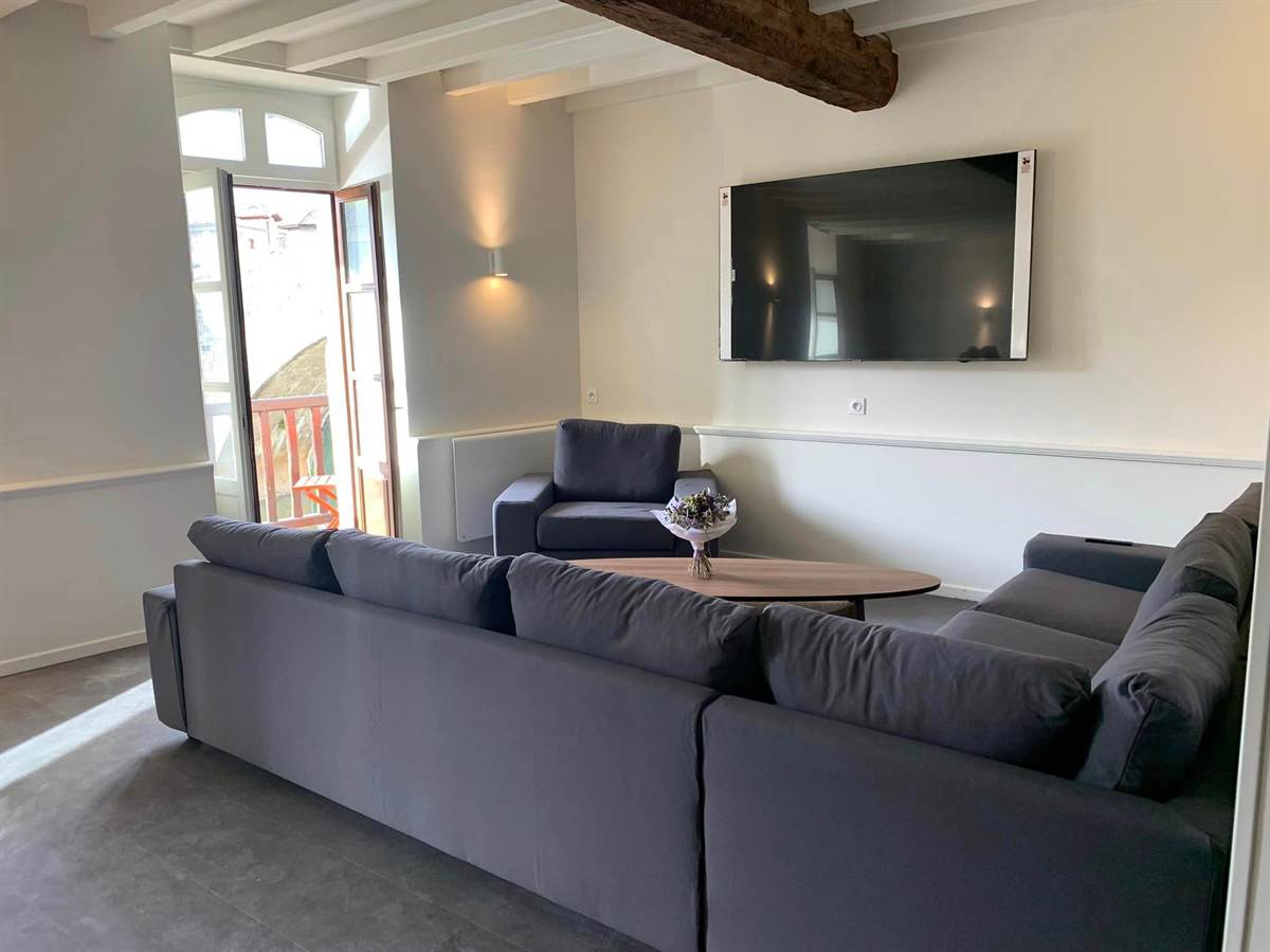 Salon avec canapé d’angle, télévision murale et porte-fenêtre sur le balcon.